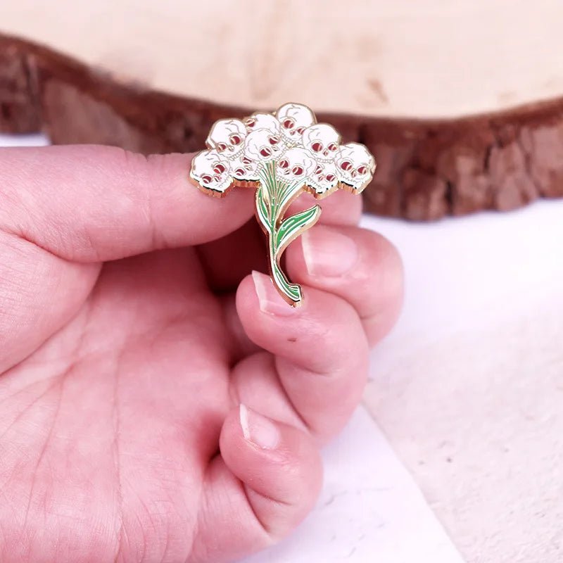 A hand holds a enamel pin shaped like a Gothic Bouquet with white flowers and green leaves, set against a blurred background on a wooden surface.