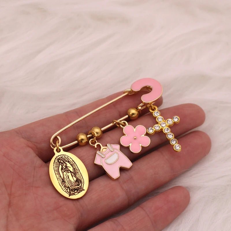 A hand holds a Kiss Me Later gold Christian baby pin brooch with four charms: Virgin Mary medallion, pink onesie, pink flower, and diamante cross with a pink question mark. The soft white fur background adds gentle elegance.