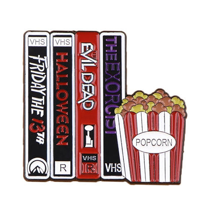 Four upright VHS tapes—Friday the 13th, Halloween, Evil Dead, The Exorcist—beside a striped “Popcorn” bucket and a spooky enamel pin, perfect for horror fans and book badge collectors.