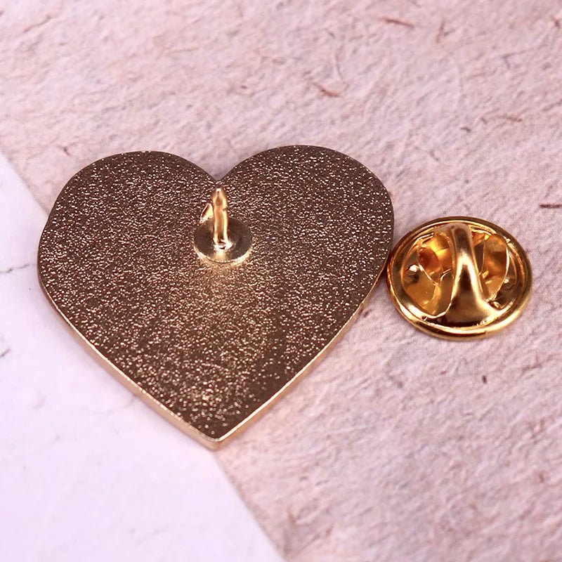  gold heart-shaped enamel pin lies face down on a textured surface beside its matching round, gold butterfly clutch backing.
