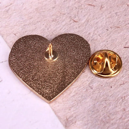  gold heart-shaped enamel pin lies face down on a textured surface beside its matching round, gold butterfly clutch backing.