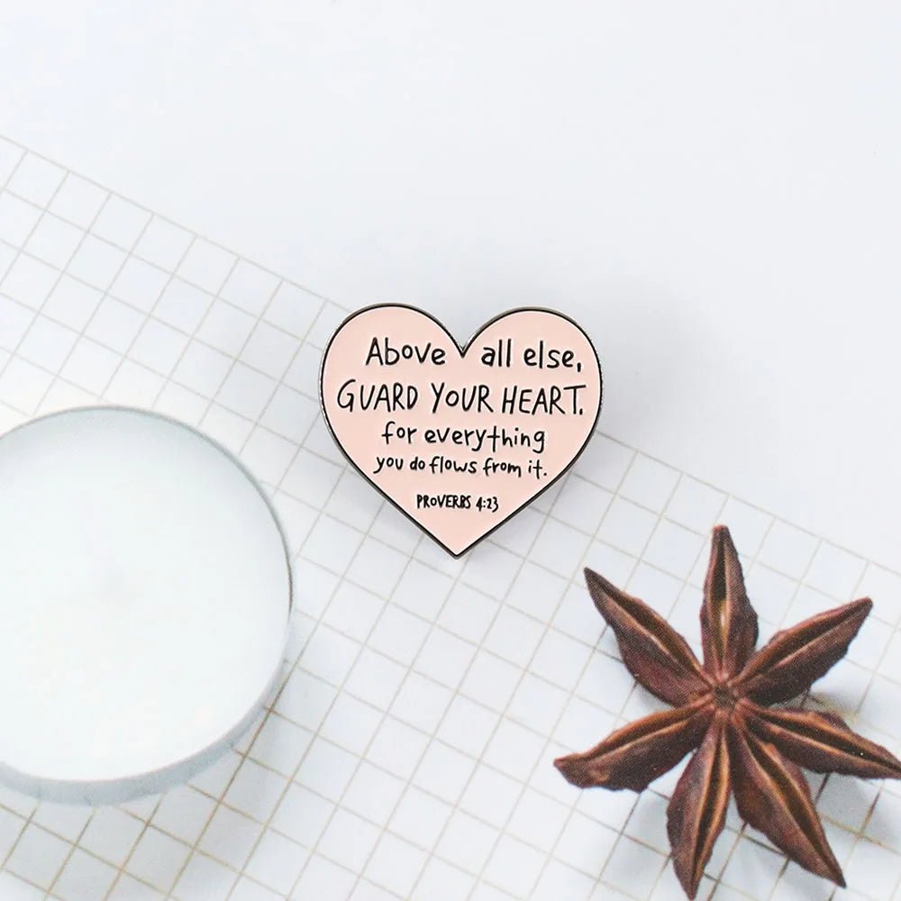 The Kiss Me Later heart-shaped enamel pin, featuring Above all else, GUARD YOUR HEART—for everything you do flows from it. Proverbs 4:23, rests on a white grid surface beside a candle and star anise.