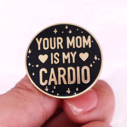 A round black enamel pin from My Cutesy Era, featuring gold Your mom is my cardio text, two heart icons, and decorative stars, held between two fingers.