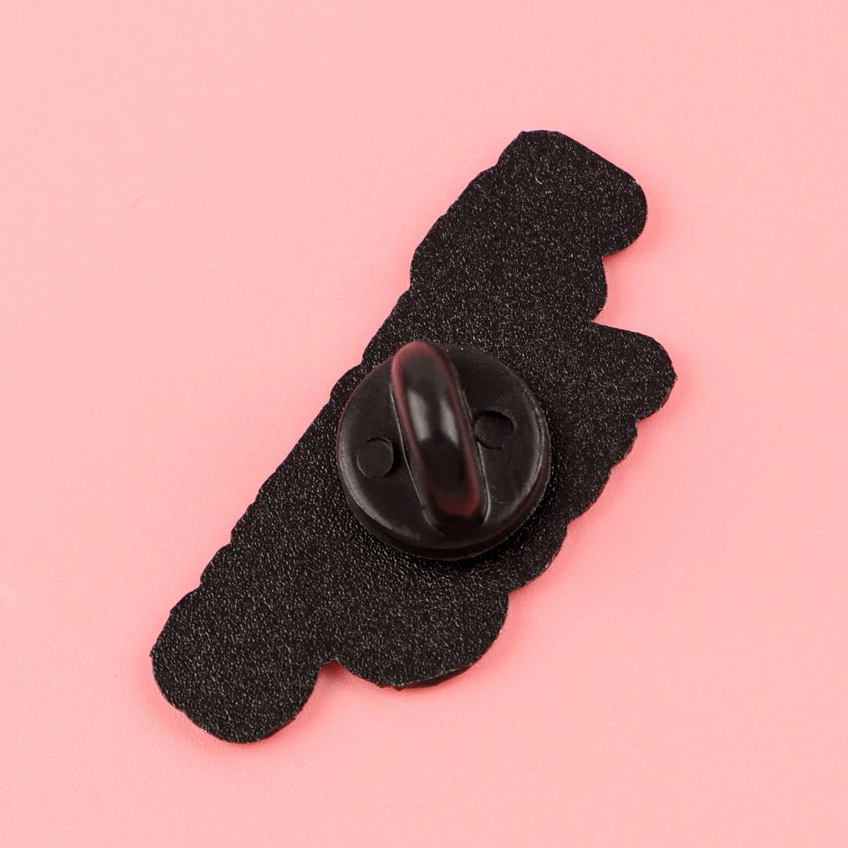 A Kiss Me Later black enamel pin backing (3.5cm x 1.5cm) with a round rubber clasp and scalloped edge is shown on a pink surface—an ideal accessory to enhance your peach pin collection.