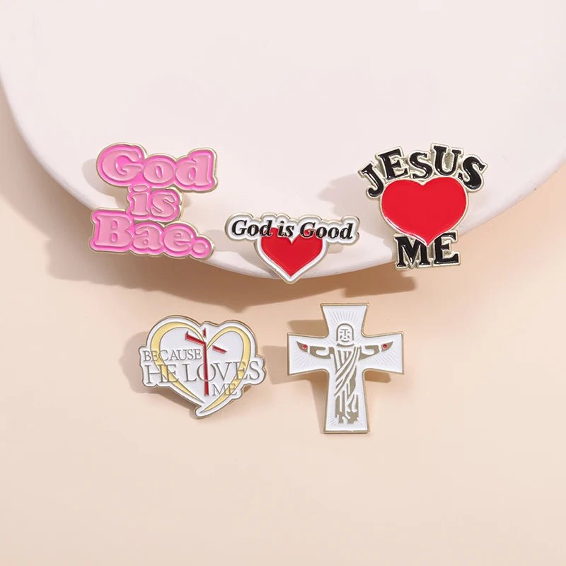Five enamel pins from Kiss Me Later on a light surface: a pink “God Is Bae” pin, “God is Good,” “JESUS ♥ ME,” “Because He Loves Me,” and a white cross with a stylized Jesus figure.