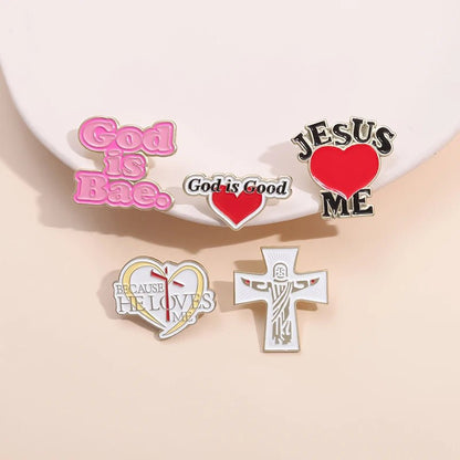 Five enamel pins from Kiss Me Later on a light surface: a pink “God Is Bae” pin, “God is Good,” “JESUS ♥ ME,” “Because He Loves Me,” and a white cross with a stylized Jesus figure.
