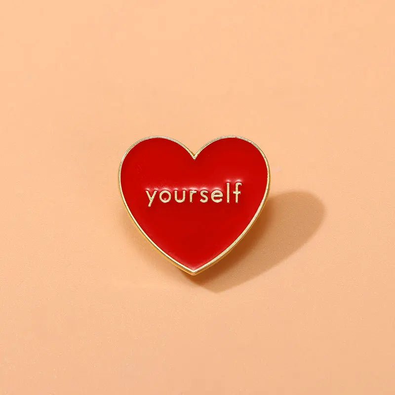 A red and gold heart-shaped enamel pin by Kiss Me Later, featuring yourself in white lowercase letters, displayed on a light peach-colored background.