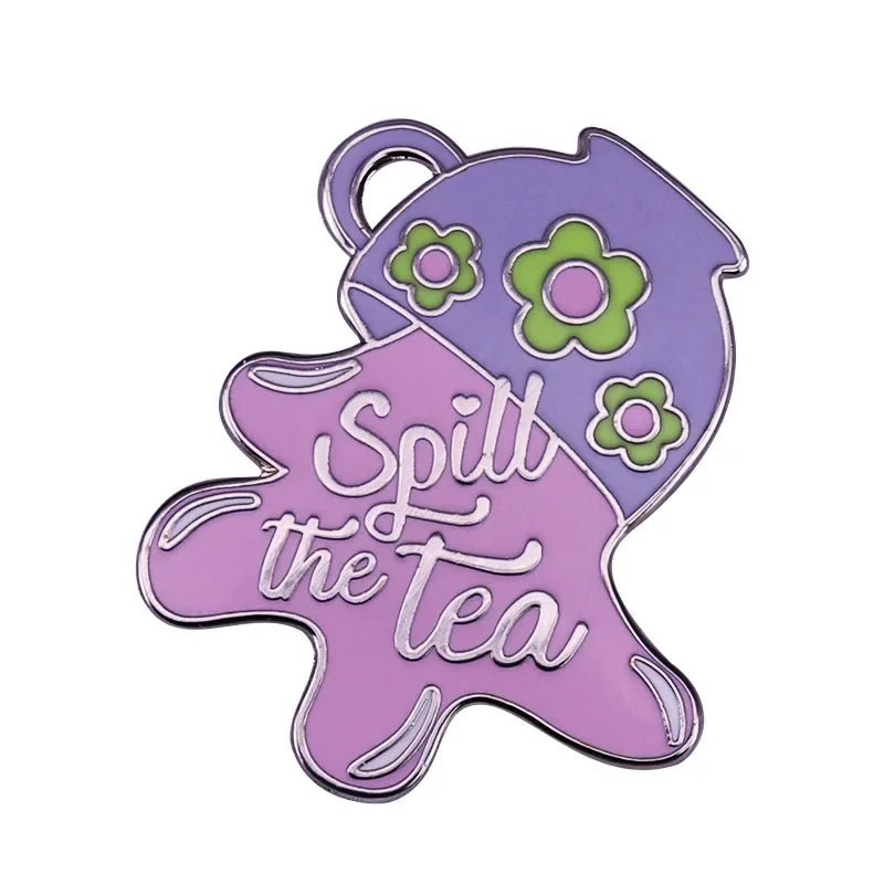 A pastel enamel pin by My Cutesy Era shaped like a tilted purple teapot with green flowers, pouring pink liquid that spells out Spill the tea in white cursive.