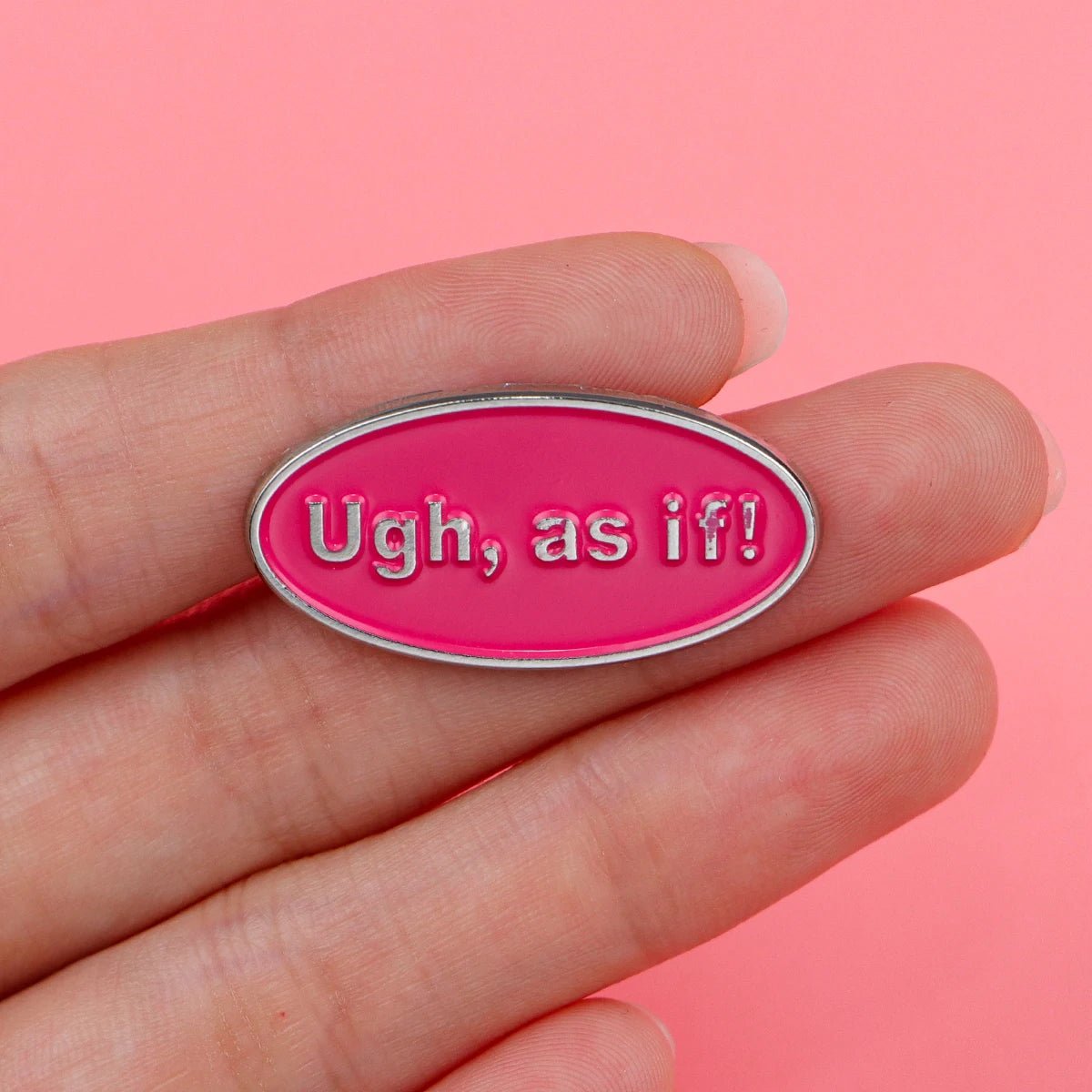 A hand holds a My Cutesy Era pink enamel oval badge with silver edges and “Ugh, as if!” embossed in white on a solid pink background.