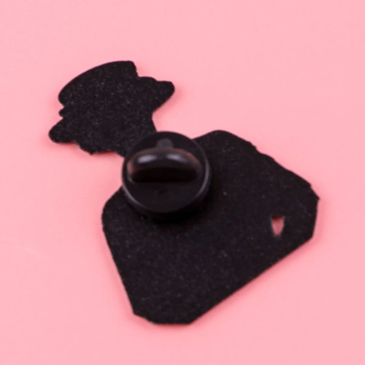 The Kiss Me Later black enamel pin shows the silhouette of a person in a hat and suit jacket, viewed from behind on a pink background. This bold feminist accessory is secured with a black rubber clutch fastener.