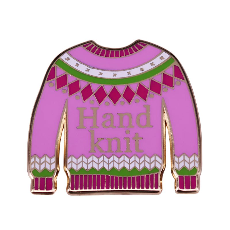 A pink enamel pin by My Cutesy Era shaped like a sweater, with green, white, and magenta patterns and “Hand knit” in gold—perfect for enamel pin collectors.