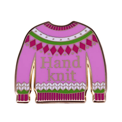 A pink enamel pin by My Cutesy Era shaped like a sweater, with green, white, and magenta patterns and “Hand knit” in gold—perfect for enamel pin collectors.