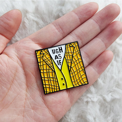 A hand holds an enamel badge, inspired by Clueless and shaped like a yellow checkered jacket over a white UGH AS IF shirt, set against a fluffy white fabric background.
