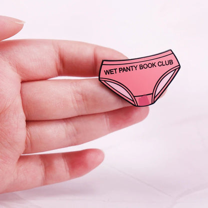 A hand displays an enamel pin shaped like pink underwear with bold WET PANTY BOOK CLUB text. The soft, light pink background highlights this playful book club accessory.
