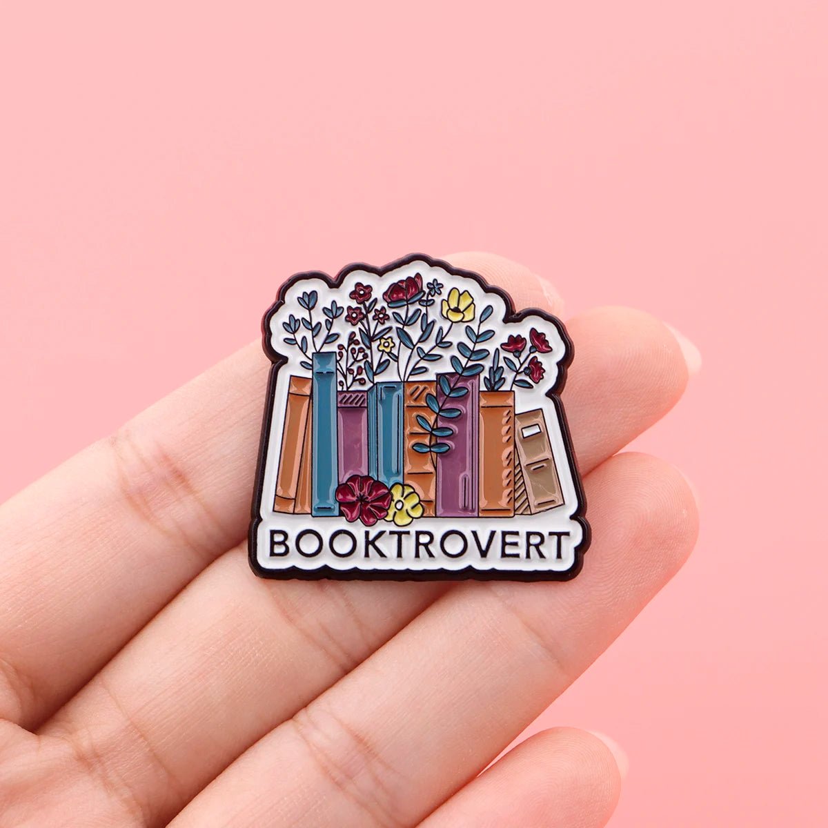 A hand holds an enamel pin with colorful books and flowers, “BOOKTROVERT” below—perfect for public library fans—set against a pink background.