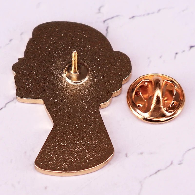 A gold lapel pin by Kiss Me Later, shaped like a womans head in profile and reminiscent of a black silhouette enamel pin, is displayed with its round backing on a white marble-like surface.