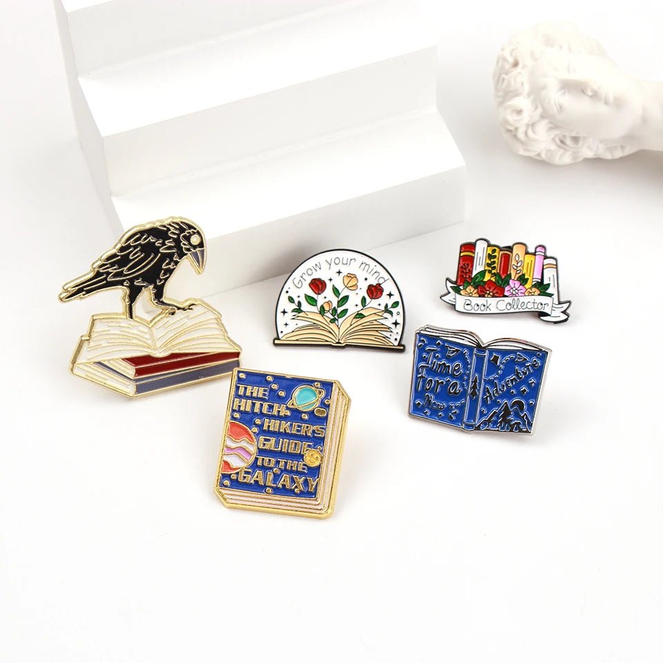 Five colorful enamel pins shaped like books, are arranged on a white surface—perfect for book lovers. Designs include a raven on books, “Grow your mind,” “Book collector,” a space book, and a blue fairy tale book.