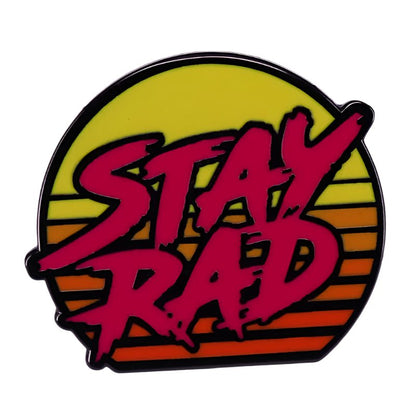 A bold, pink STAY RAD enamel pin by My Cutesy Era features stylized text over a yellow-orange sunset with horizontal stripes—perfect for pin collectors or fans of unique accessories.