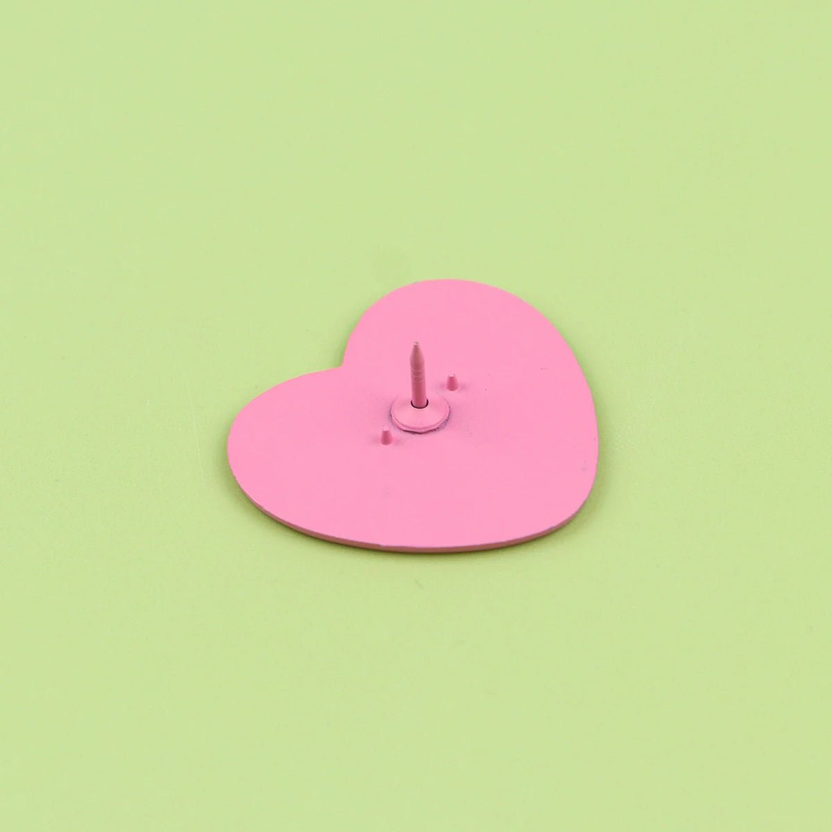 A Kiss Me Later pink love heart pin with a small sharp spike in its center is displayed on a light green background.