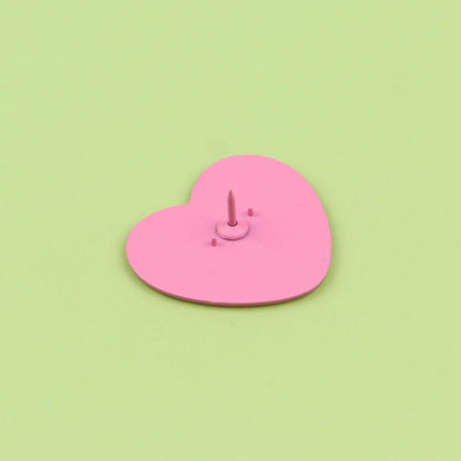 A Kiss Me Later pink love heart pin with a small sharp spike in its center is displayed on a light green background.