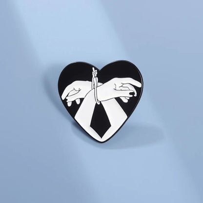 The Kiss Me Later Bound Hands enamel pin features two illustrated hands tied at the wrists in a love heart shape, set against a black background and displayed on a light blue surface.
