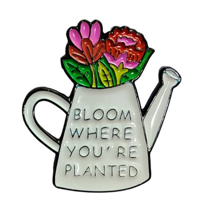 A My Cutesy Era enamel pin featuring a white watering can with vibrant flowers, black and gold accents, and the phrase BLOOM WHERE YOU’RE PLANTED—a perfect accessory for the Black Sheep Club.