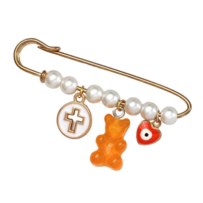 The Kiss Me Later gold safety pin brooch features white pearl beads and three charms: a gold cross in a white circle, an orange gummy bear, and a red heart with an eye—perfect as a unique baby shower gift.