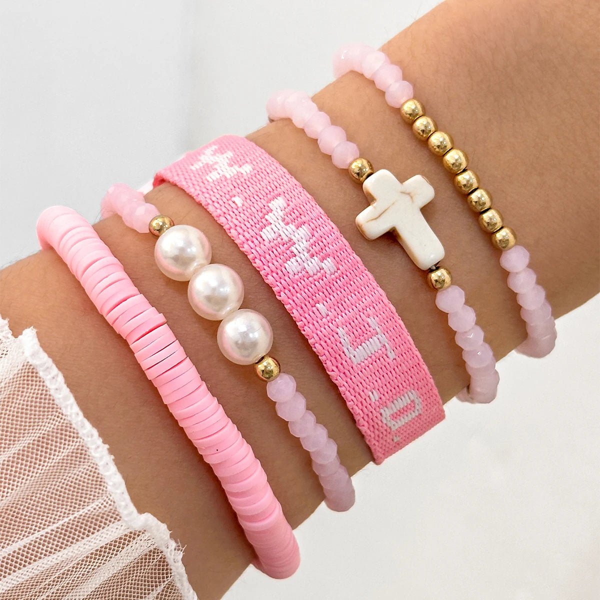 A wrist styled with a Kiss Me Later pink bracelet stack—five pink and gold beaded bracelets with pearls, a white cross, and a woven bracelet featuring white text—worn above a sheer white ruffled sleeve.