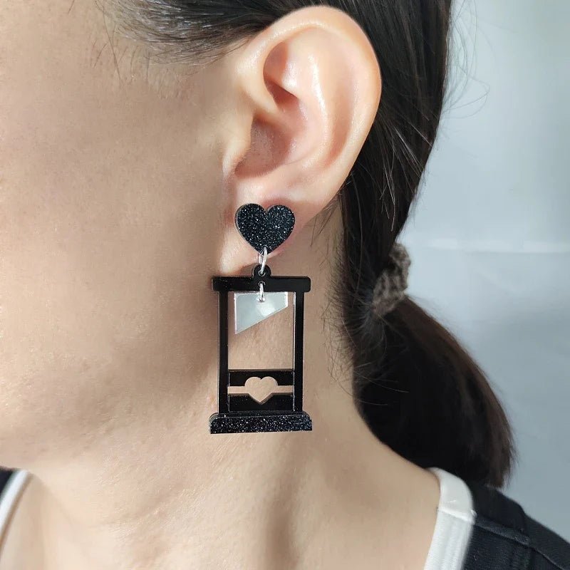 A close-up of a person’s ear wearing Gay Skate guillotine acrylic earrings with black hearts at the top and small hearts at the base. Their hair is tied back with a brown scrunchie, letting the bold pink and black earrings stand out.