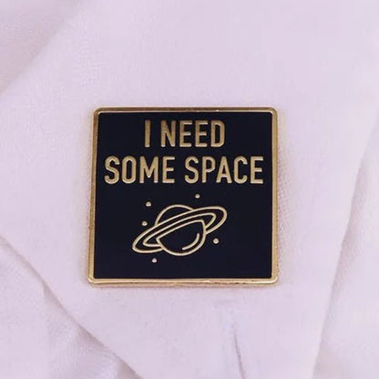 A square black and gold metal brooch by Kiss Me Later reads “I NEED SOME SPACE” above a planet with rings—an on-trend star pin perfect for any unisex accessory collection.