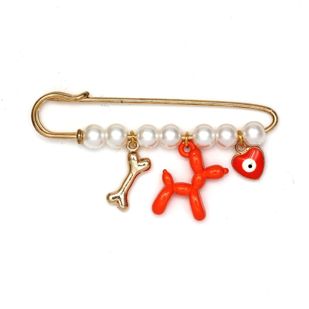 The Kiss Me Later gold safety pin brooch features five white pearls and three charms—a gold dog bone, an orange balloon dog, and a red heart with an eye—making it a unique, lovely gift for a baby shower.