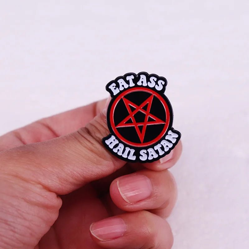 A hand holds a Kiss Me Later enamel pin with a red pentagram in the center and EAT ASS HAIL SATAN in white letters on a black background.