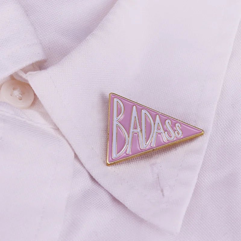 A bold  pink triangular enamel pin with BADASS in white and gold is pinned to a white shirt collar—a feminist accessory showing well-behaved women rarely make history.