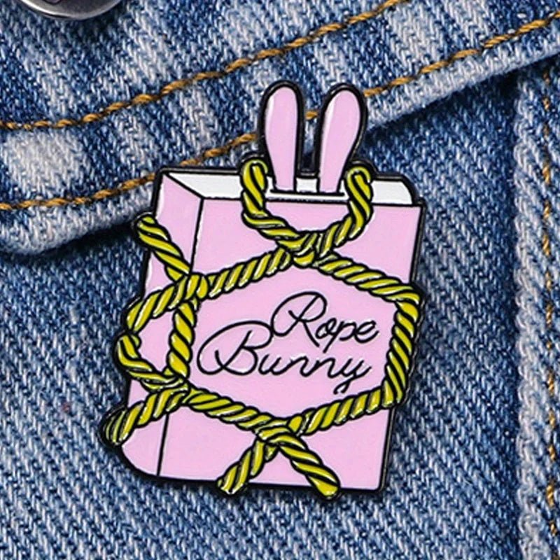 Pink rope bunny enamel pin is shaped like a book with bunny ears, wrapped in yellow rope and Rope Bunny on the cover—a playful nod to bondage style, shown pinned to blue denim fabric.