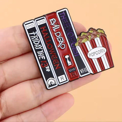 A hand holds an enamel pin shaped like VHS tapes of classic horror films and popcorn—perfect for book lover badges—against a peach background.