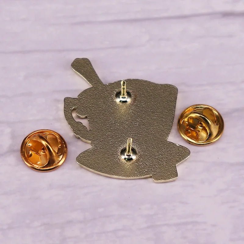 A My Cutesy Era metallic enamel pin with two posts and gold butterfly clasps is shown facing backwards on a light purple surface. Its irregular shape suggests a floral teacup or whimsical character outline.