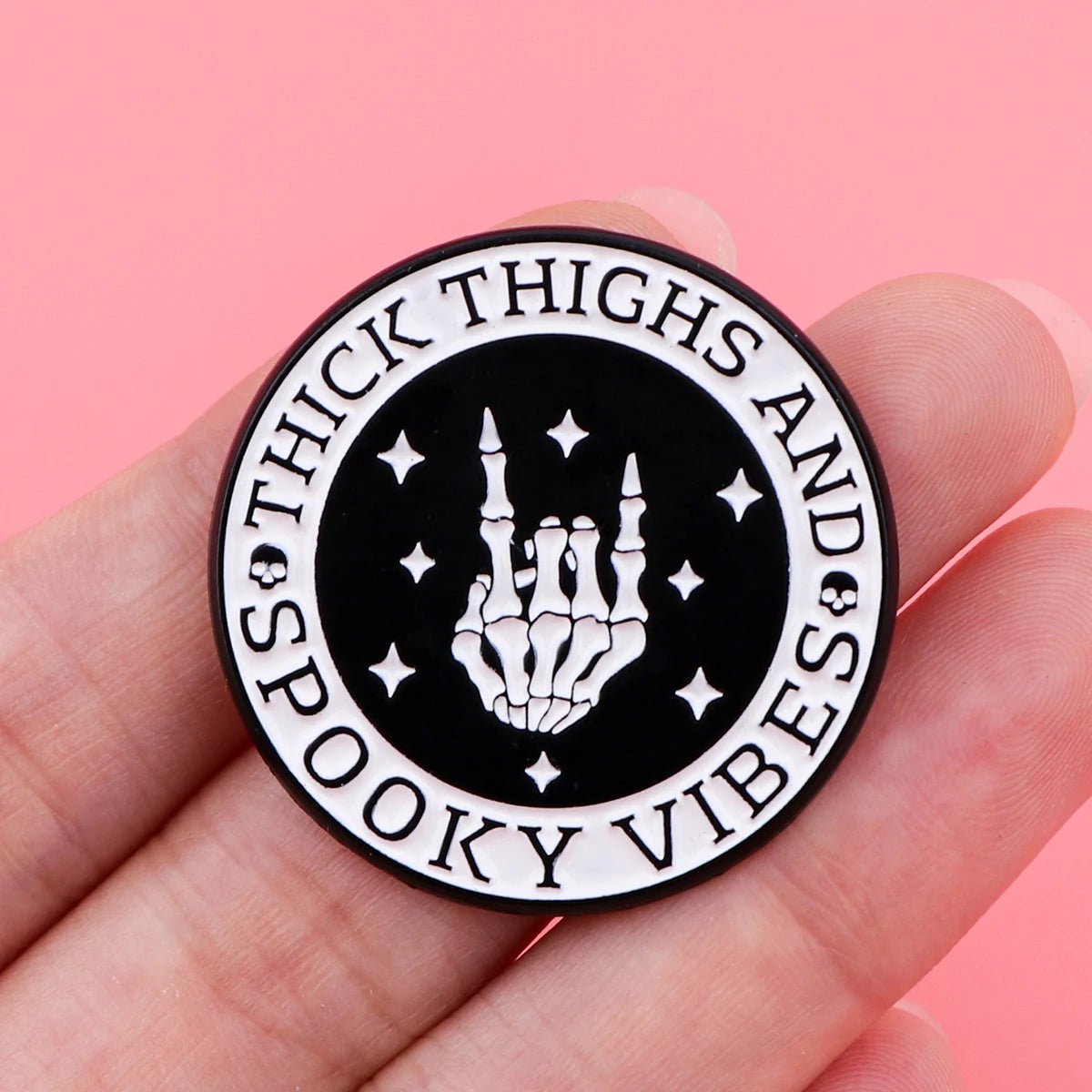 A round black and white enamel pin by Kiss Me Later features a skeleton hand flashing the rock on sign, stars, and “THICK THIGHS AND SPOOKY VIBES” along the edge—perfectly popping against a bold pink background.