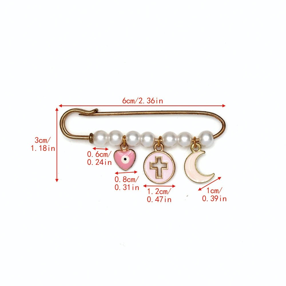 The Kiss Me Later gold safety pin brooch features white beads and four dangling charms—a pink heart, a cross in a circle, and a crescent moon—making it a lovely baby shower gift. Red labels show charm and pin lengths in cm and inches.