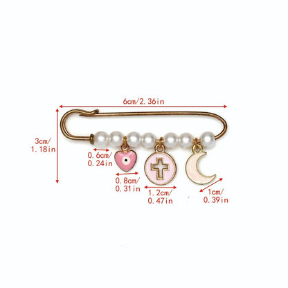 The Kiss Me Later gold safety pin brooch features white beads and four dangling charms—a pink heart, a cross in a circle, and a crescent moon—making it a lovely baby shower gift. Red labels show charm and pin lengths in cm and inches.