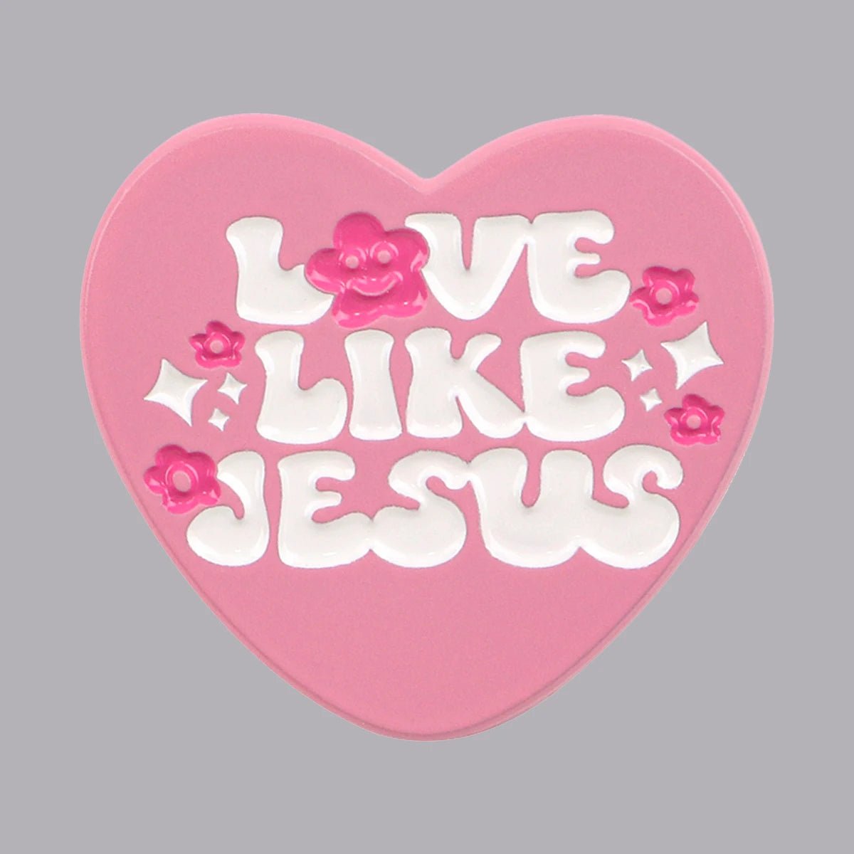 The Kiss Me Later pink love heart pin features white, bubbly “Love Like Jesus” lettering, accented with small pink flowers and sparkles, shining brightly against a gray background.