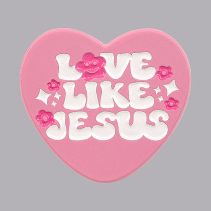 The Kiss Me Later pink love heart pin features white, bubbly “Love Like Jesus” lettering, accented with small pink flowers and sparkles, shining brightly against a gray background.
