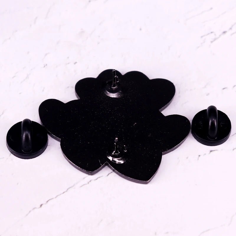 A black flower-shaped enamel pin by featuring two detached black rubber pin backs, displayed on a white marble surface—the perfect Book Lover accessory for any reader’s collection.