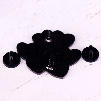 A black flower-shaped enamel pin by featuring two detached black rubber pin backs, displayed on a white marble surface—the perfect Book Lover accessory for any reader’s collection.
