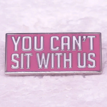 A rectangular pink enamel pin features white and silver text reading YOU CANT SIT WITH US—the perfect accessory for Mean Girls fans, shown on a light textured surface.