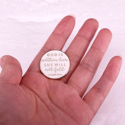 A hand holds a white and gold enamel pin engraved with “God is within her, she will not fall. Psalm 46:5” against a pale background.