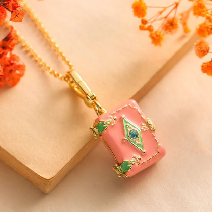 Gold pink enamel necklace featuring a suitcase-shaped charm with green and gold detailing and a blue gem is displayed on beige, accompanied by orange dried flowers.