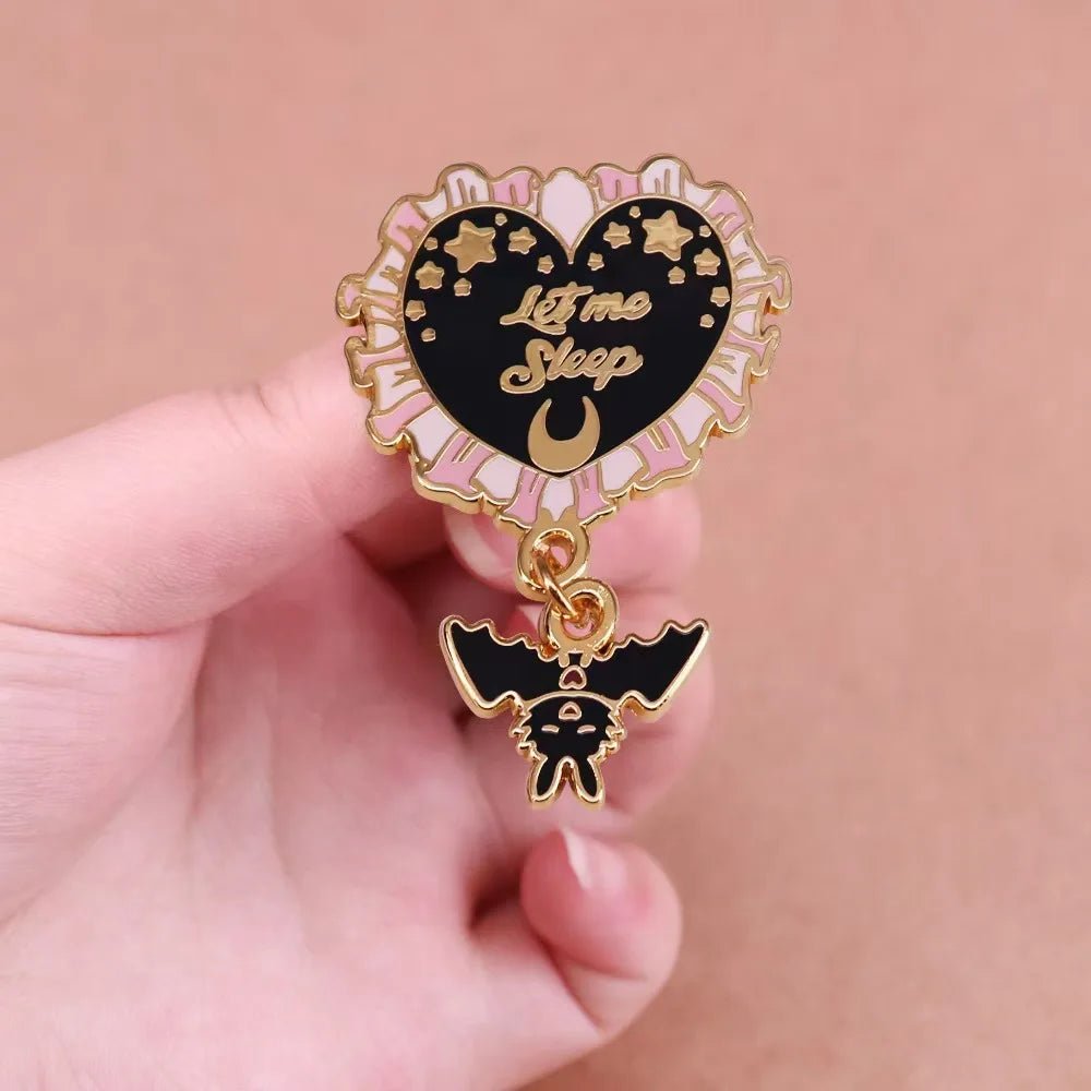 A hand holds an enamel pin: pink and black with a black heart, stars, gold Let Me Sleep text, pink frills, and a dangling bat charm—ideal for any heart or bat-themed pin collection.