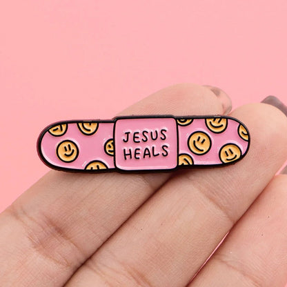A hand holds a pink bandaid enamel pin with yellow smiley faces and JESUS HEALS in the center, set against a solid pink background.