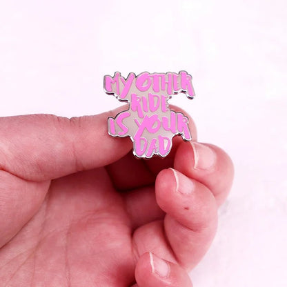 A hand holds an enamel pin with bold text, MY OTHER RIDE IS YOUR DAD, against a light pink background.