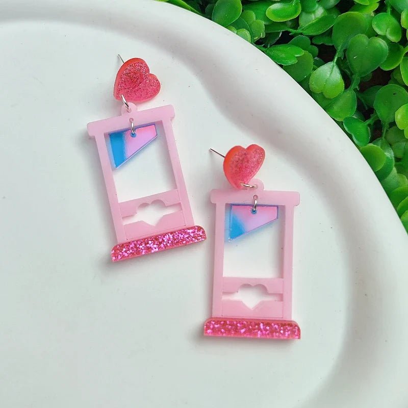 Kiss Me Later’s sparkly acrylic earrings feature pink window shapes with blue and white panes, heart cutouts, glittery pink bases, and red glitter heart studs, displayed on a white surface with green leaves in the background.